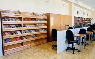 Rząd stolik&oacute;w z krzesłami wypełnia środek pomieszczenia, obok, pod ścianą regały z czasopismami i książkami