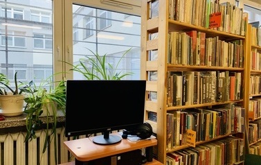 Biurko z komputerem stojące przy oknie, obok regał z książkami