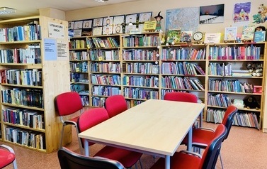 Na środku pomieszczenia st&oacute;ł z sześcioma czerwonymi krzesłami, dookoła niego regały z książkami i wystawa prac plastycznych