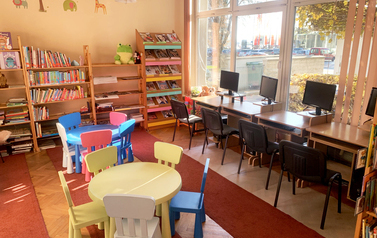 Sala z regałami z książkami i stanowiskami komputerowymi, na środku kolorowe stoliki i krzesełka dla dzieci