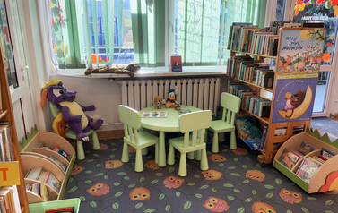 Kącik dla dzieci z p&oacute;łkami z książkami, zielonym stoliczkiem z krzesełkami i szarym dywanem w pomarańczowe sowy
