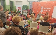 W sali zgromadzone osoby siedzą na spotkaniu autorskim, kobieta trzyma mikrofon i podstawia drugiej kobiecie do buzi. Obok siedzi mężczyzna.