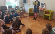 Kobieta stoi i mówi do dzieci siedzących na podłodze.