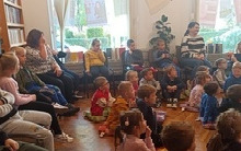 Kilkanaście osób siedzi na podłodze przed nimi kobieta stojąca.