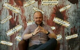 Wojciech Chmielarz siedzi na krześle, w tle tytuły jego książek. 
