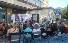 Na krzesłach siedzą rodzice z dziećmi przed budynkiem.