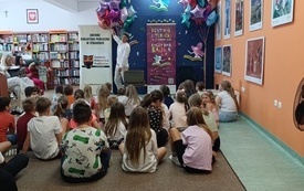 Opowiadacz prowadzi zajęcia dla dzieci zebranych w bibliotece. W tle kolorowe balony. 