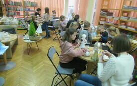 Grupa dzieci i dorosłych siedzi przy stolikach, na kt&oacute;rych leżą r&oacute;żne materiały plastyczne