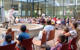 Na dziedzińcu spora grupa dzieci. Mężczyzna w kapeluszu stoi na fontannie i pokazuje coś. 