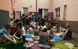 Na dziedzińcu siedzą dzieci i słuchają kobiety opowiadającej. 
