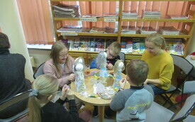 Dzieci i dorosłą kobieta siedzą przy stoliku i tworzą figurki anioł&oacute;w