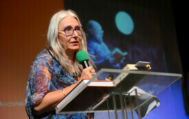 Kobieta w siwych włosach i okularach przemawia na scenie do mikrofonu.