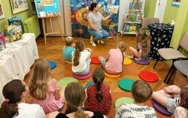 W bibliotece na podłodze siedzą dzieci i słuchają opowiadań bibliotekarki. 