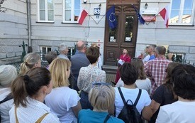 Kobieta w r&oacute;żowej sukience z mikrofonem stoi wraz z uczestnikami spaceru przed budynkiem Uniwersytetu Rzeszowskiego.