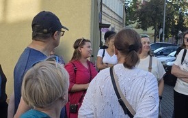 Grupa ludzi zebranych wok&oacute;ł kobiety w r&oacute;żowej sukience z mikrofonem.
