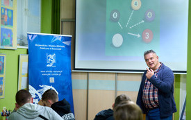 Prowadzący spotkanie mężczyzna m&oacute;wi do zgromadzonej publiczności. W tle prezentacja multimedialna