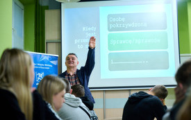 Prowadzący spotkanie mężczyzna z podniesioną do g&oacute;ry ręką m&oacute;wi do zgromadzonej publiczności. W tle prezentacja multimedialna.