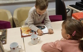 Dziecko przy stoliku czyta książkę, a dziewczynka naprzeciwko niego słucha