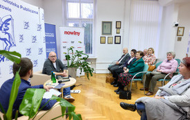 Rozmowa dwóch mężczyzn, po prawej zebrana publiczność.