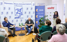 Pisarz, prowadzący spotkanie oraz bibliotekarka są zwróceni twrzą do publiczności.