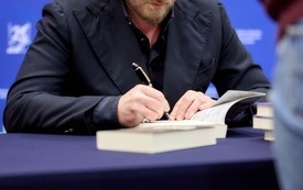 Pisarz w okularach siedzący przy stoliku i podpisujący książki. 