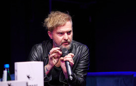 Mężczyzna z bujną czupryną w czarnej sk&oacute;rze i podkoszulku z mikrofonem w ręku.