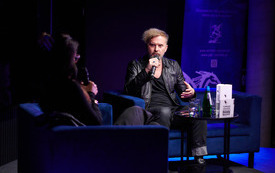 Mężczyzna w czarnej sk&oacute;rze i podkoszulku z mikrofonem w ręku, obok prowadząca spotkanie kobieta.