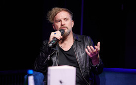 Mężczyzna w czarnej sk&oacute;rze i podkoszulku z mikrofonem w ręku.