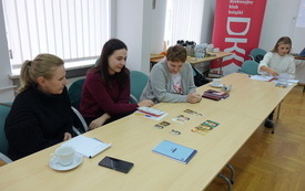 Trzy kobiety patrzą na ułożone na stole zdjęcia. W tle stolik i baner DKK.