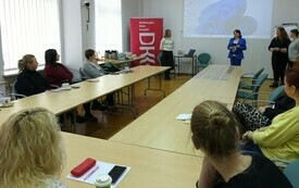 Przy stole siedzą uczestnicy spotkania. Przed nimi stoją cztery kobiety. 