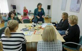 Przy dw&oacute;ch stolikach uczestnicy spotkania. Przy każdym ze stolik&oacute;w prowadząca spotkanie kobieta