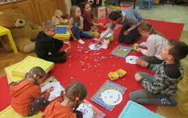 Dzieci siedzą na poduszkach i dekorują papierową bombkę, a prowadząca rozdaje elementy do dekoracji 