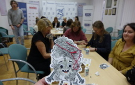 Stworek z papieru z włosami z wł&oacute;czki. W tle uczestnicy warsztat&oacute;w.