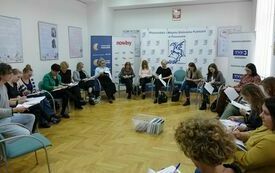 Pośrodku sali pudełko z książkami, wok&oacute;ł uczestnicy spotkania piszący coś na kartkach.