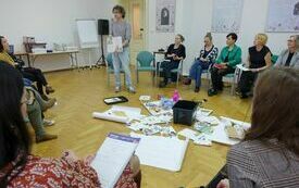 Prowadzący spotkanie z książką w ręku zwr&oacute;cony do siedzących wok&oacute;ł uczestnik&oacute;w warsztat&oacute;w.