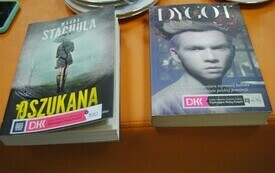 Na stole leżą dwie książki. Pierwsza z nich przedstawia kobietę w płaszczu, druga twarz młodego mężczyzny