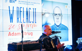 Mężczyzna podczas występu, w tle grafika informująca o koncercie. 