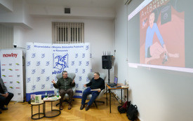 Autor i prowadzący rozmawiają gestykulując, na ścianie widoczna prezentacja ilustracji