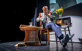 Mężczyzna siedzi na krześle na scenie, obok niego wiklinowy stolik i stare radio będące częścią scenografii do spektaklu