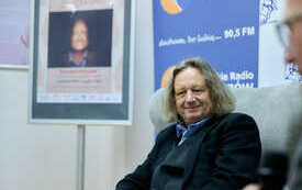 Mężczyzna z kręconymi włosami siedzący w fotelu. Obok plakat z jego zdjęciem.