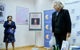 Kobieta z mikrofonem przedstawia gościa spotkania, starszego mężczyzną w szpakowatych włosach.