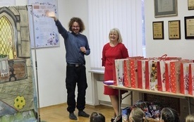 Grupa dzieci siedzi na podłodze, przed nimi mężczyzna z dłuższymi włosami o kobieta w czerwonej sukience. 