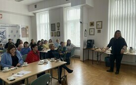 Grupa kobiet siedzi przy stolikach w sali. Przed nimi stoi prowadząca spotkanie kobieta.