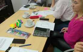 Grupa kobiet siedzi przy stoliku. Na stoliku notesy, filiżanki, flamastry