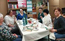 Grupa kobiet siedzi przy stole, jedna z nich na końcu stołu śmieje się, a pozostałe spoglądają na nią