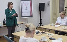 Prowadząca spotkanie kobieta, obok przy stolikach uczestnicy.