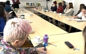 Przy stole grupa os&oacute;b, przed nimi kobieta z książką w ręku.