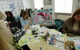 Kobieta trzyma w ręku prace plastyczną. W tle inni uczestnicy warsztat&oacute;w podczas pracy. 