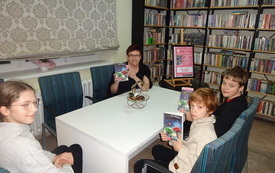 Troje dzieci, zwr&oacute;conych przodem, siedzi przy stole, a na drugim planie kobieta prezentuje ksiązkę.