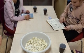 Dwie dziewczynki siedzą przy stole, na pierwszym planie znajduje się miska z popcornem. Jedna z dziewczynek czyta książkę, a druga patrzy przed siebie.